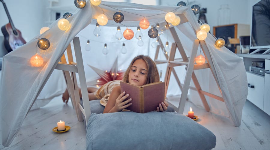 Niña leyendo libro en fuerte de mantas iluminado, aprendizaje creativo con Berlitz.