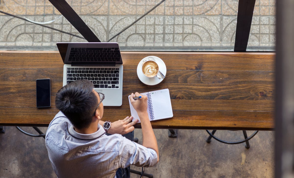 Un hombre joven concentrado, escribiendo en su libreta junto a su laptop y un café.