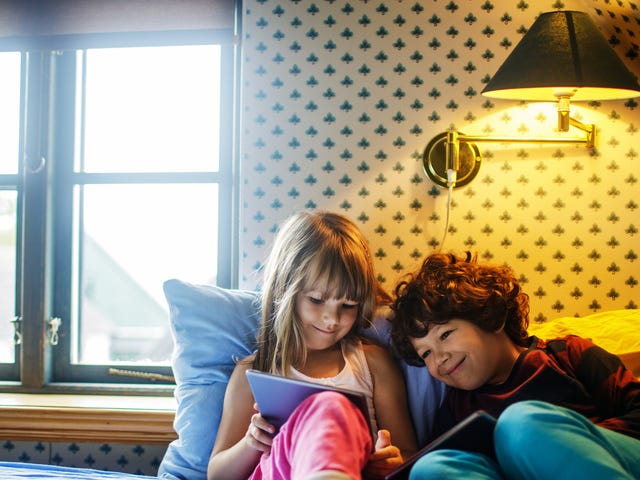 Dos niños cómodamente recostados en la cama, entretenidos con sus tablets.