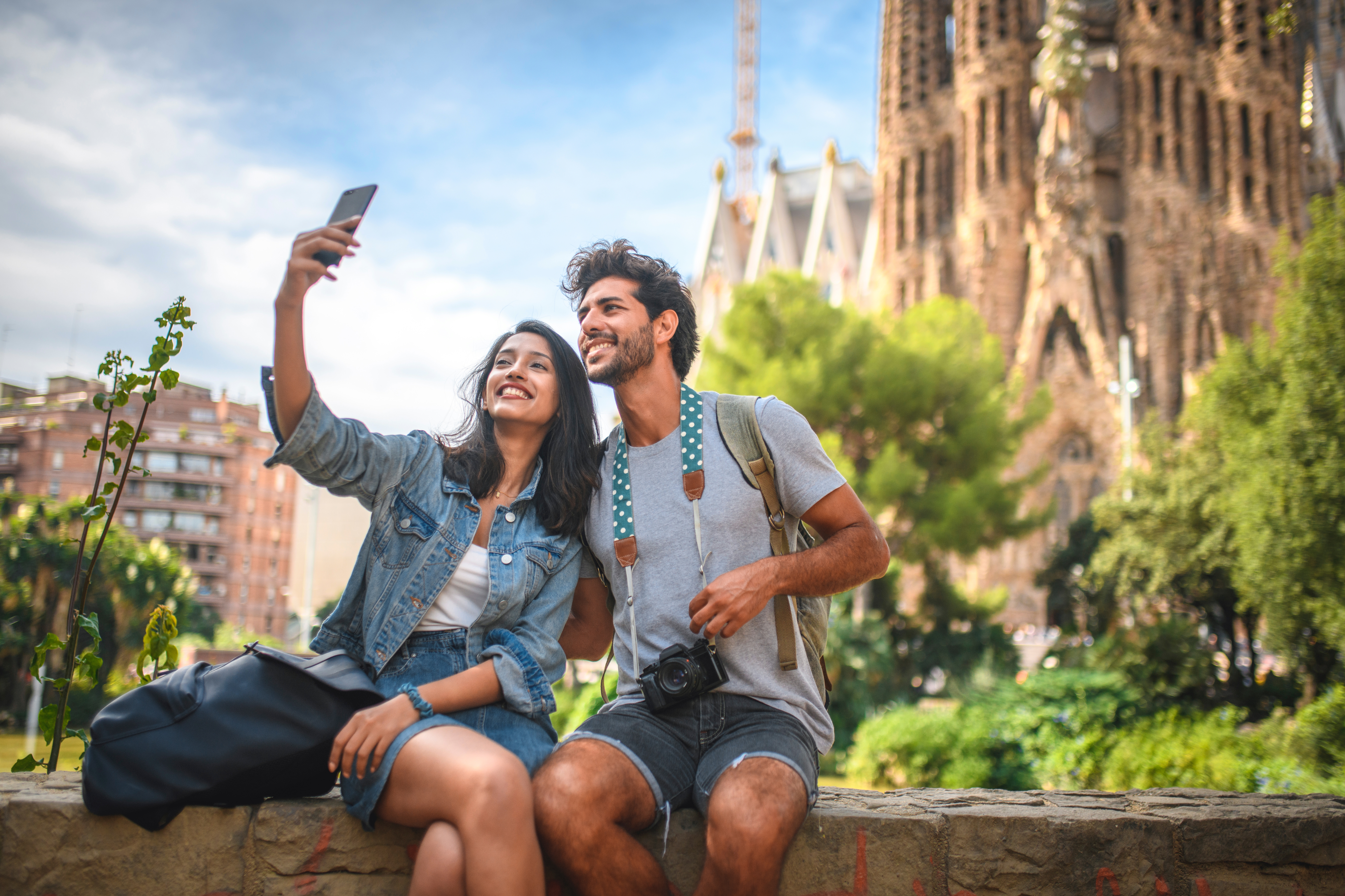 Una pareja joven y feliz se toma una selfie frente a un edificio histórico en un día soleado.