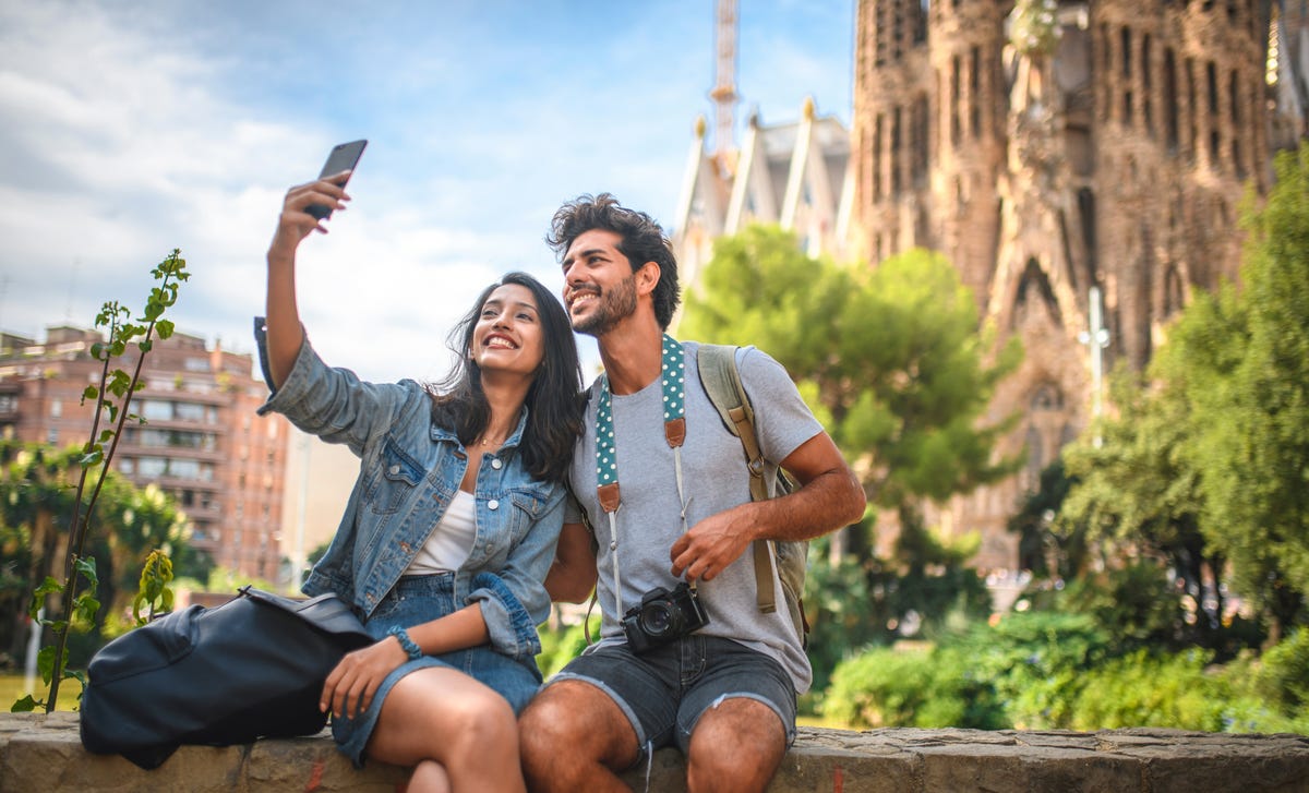 Una pareja joven y feliz se toma una selfie frente a un edificio histórico en un día soleado.