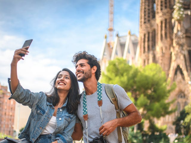 Una pareja joven y feliz se toma una selfie frente a un edificio histórico en un día soleado.