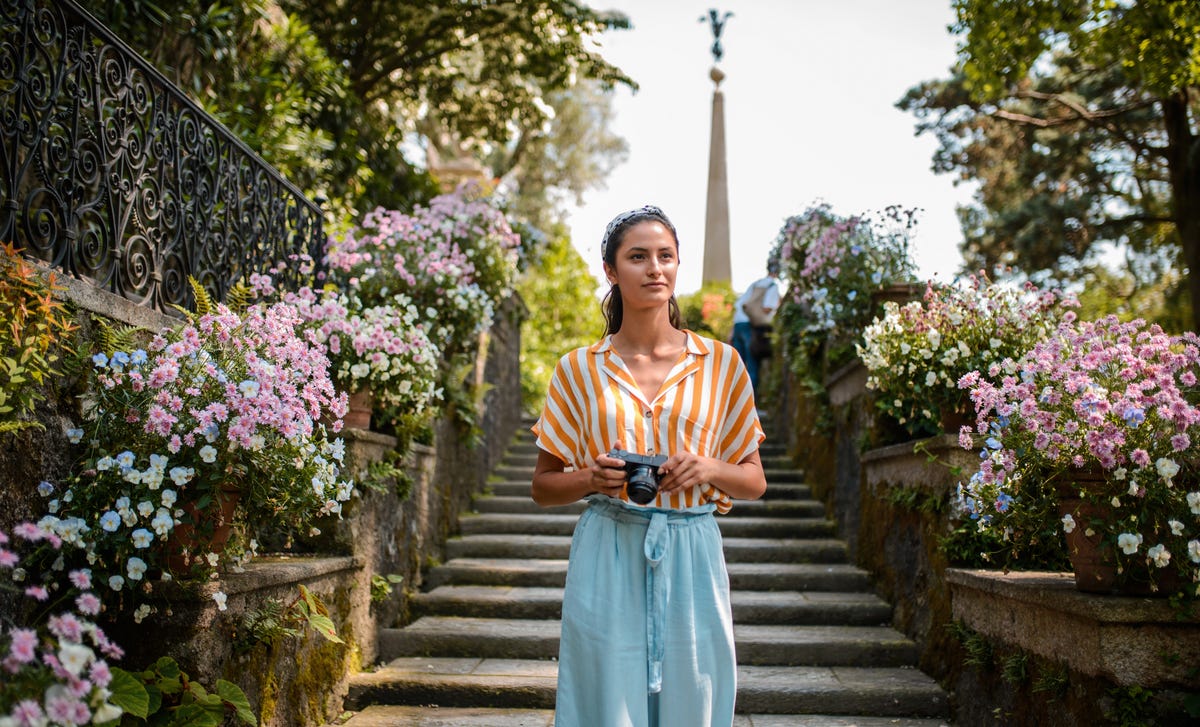 Una joven turista sostiene una cámara mientras sube una escalera exterior rodeada de flores.