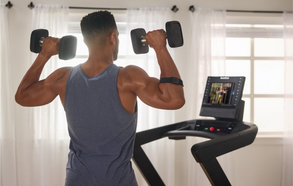 Individual performing a dumbbell shoulder press while following a guided iFIT workout on a NordicTrack treadmill screen at home.