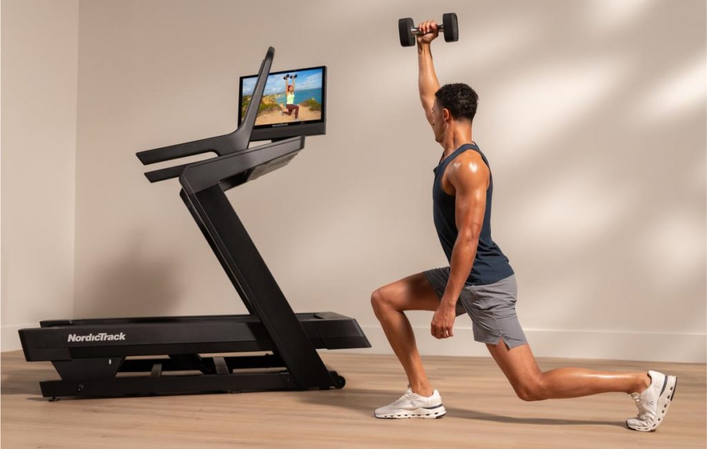 Individual performing a weighted lunge while following an iFIT strength workout on a NordicTrack treadmill screen at home.