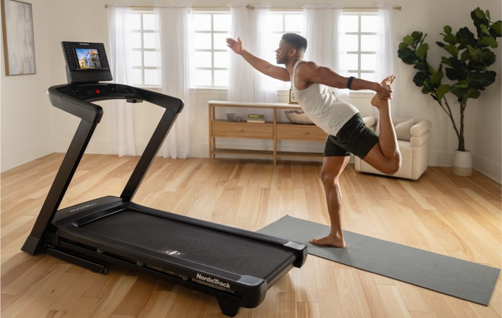 Individual performing a balance stretch beside a NordicTrack treadmill during a low-intensity recovery session at home.
