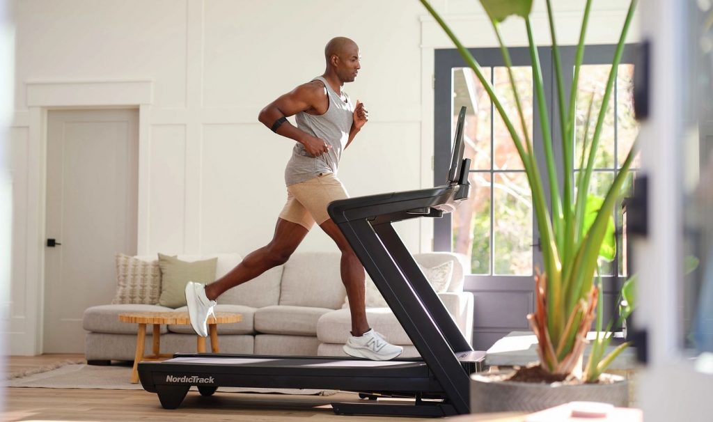Runner training on a NordicTrack treadmill in a bright living space, showcasing performance capability, cushioning, and immersive screen experience.