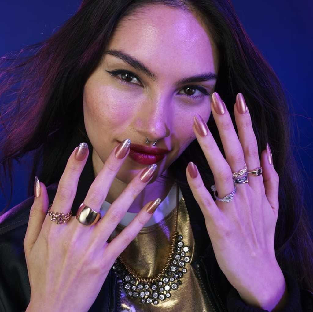 Model showcasing metallic nude nail polish with long nails, wearing rings and a statement necklace.