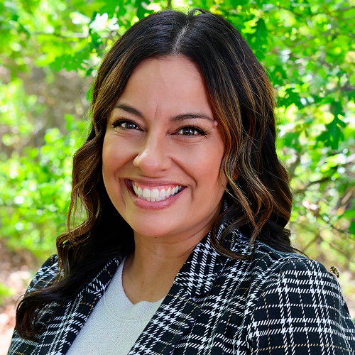 Individual with dark hair wearing a checked jacket, standing in front of a leafy green background. Head and face are centered but blurred out.