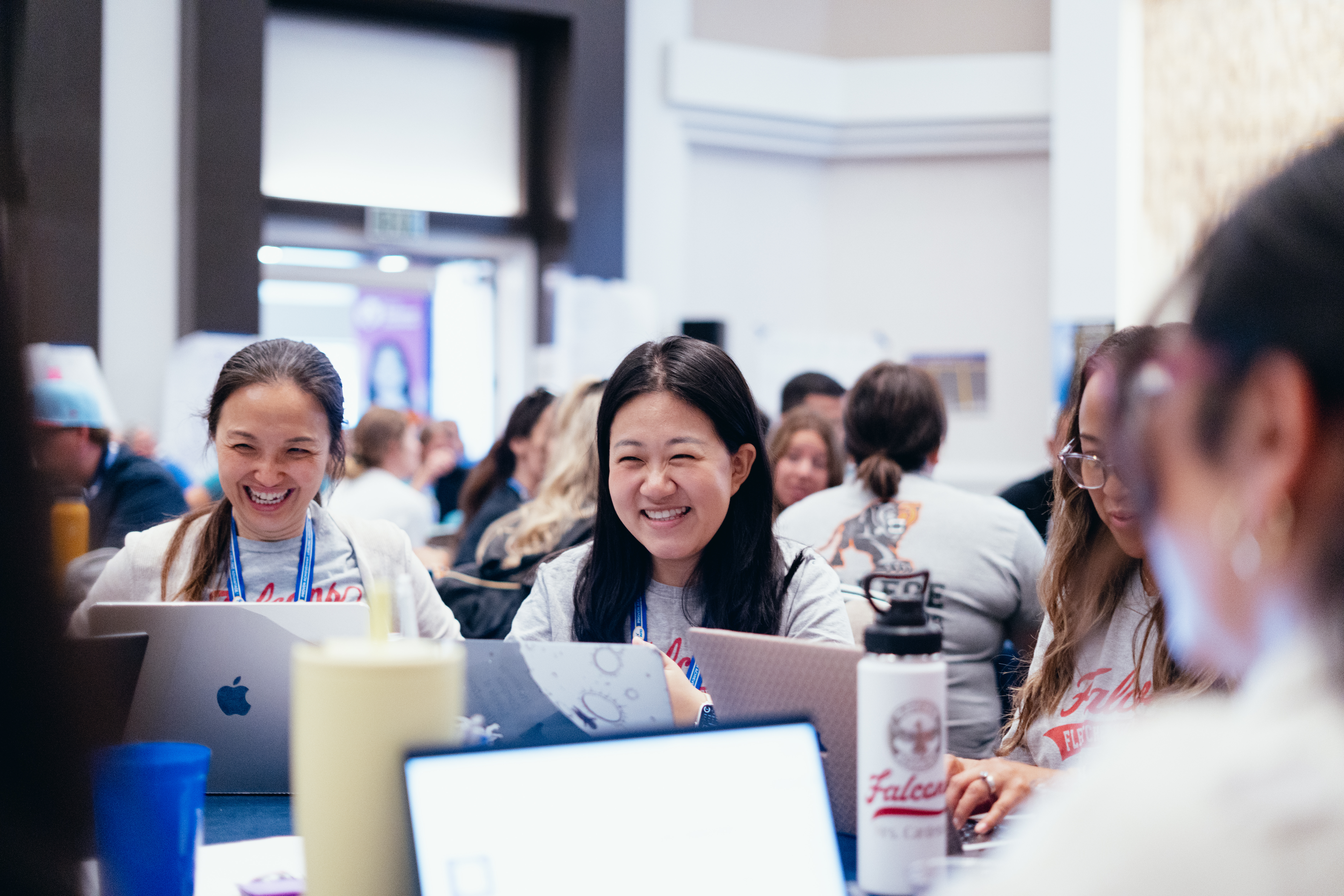 Group of people in a brightly lit room, sitting at tables with laptops and various drink containers.