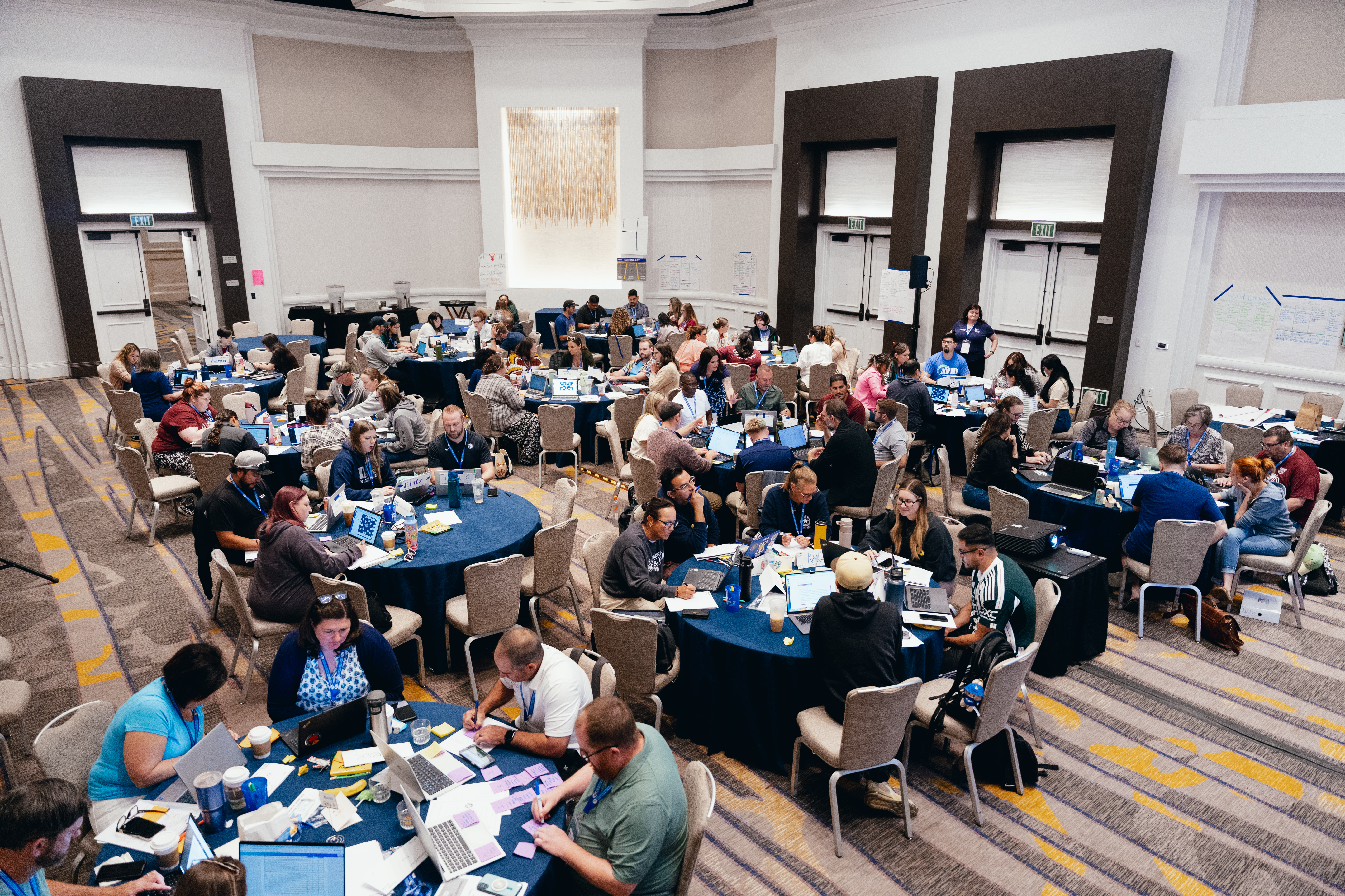 A large group of people sitting at round tables, working on laptops and discussing in a spacious conference room.