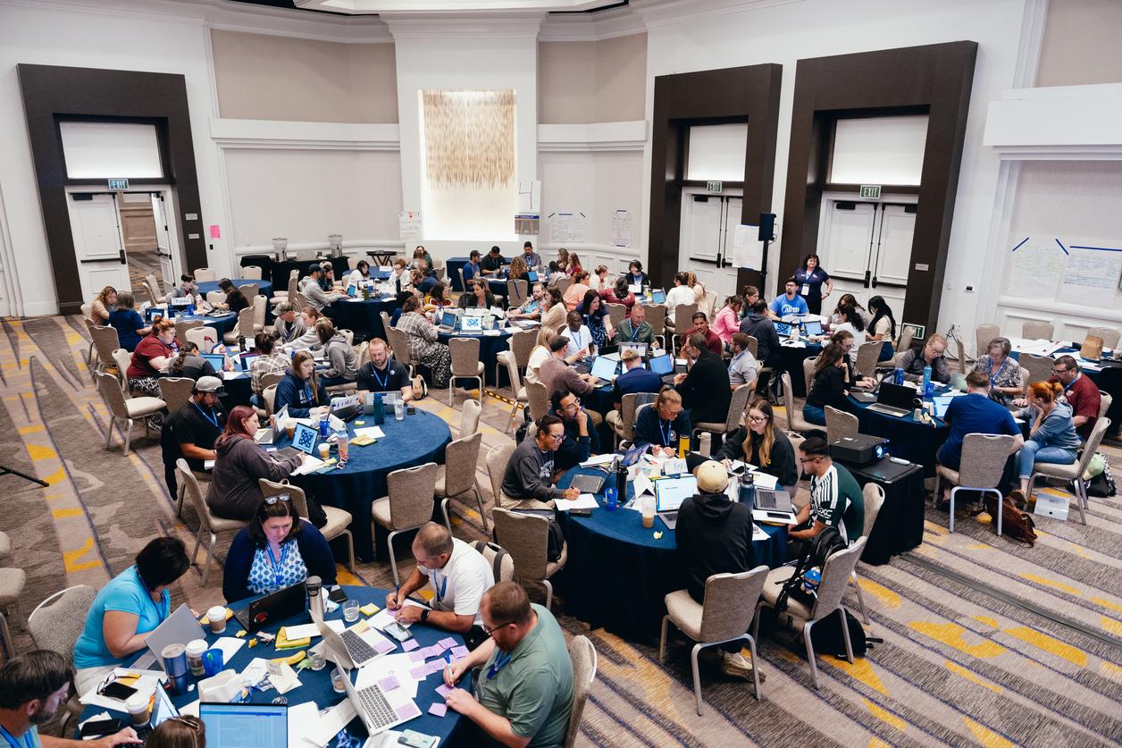 A large group of people sitting at round tables, working on laptops and discussing in a spacious conference room. A large group of people sitting at round tables, working on laptops and discussing in a spacious conference room.