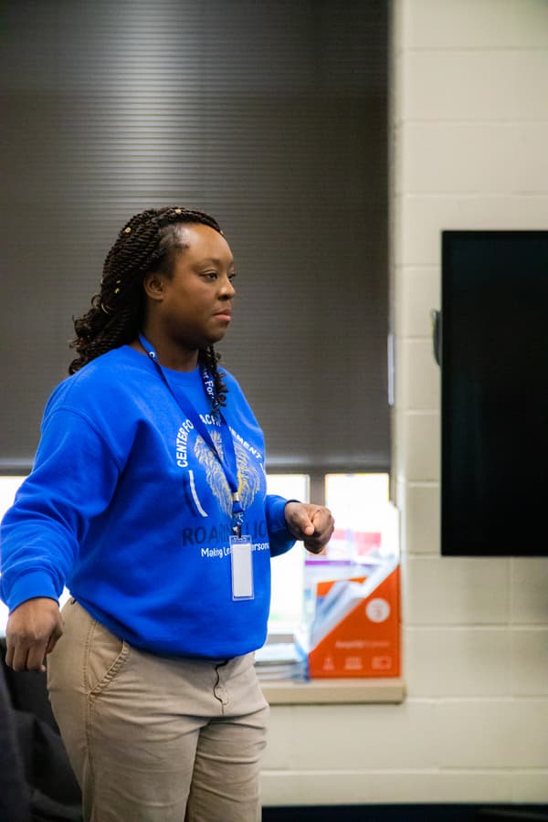 A person standing indoors wearing a blue sweatshirt and khaki pants, with a badge lanyard around the neck. The background features a window and a television screen. A person standing indoors wearing a blue sweatshirt and khaki pants, with a badge lanyard around the neck. The background features a window and a television screen.