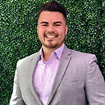 Person wearing a light grey suit jacket and lavender shirt standing in front of a green leafy background.