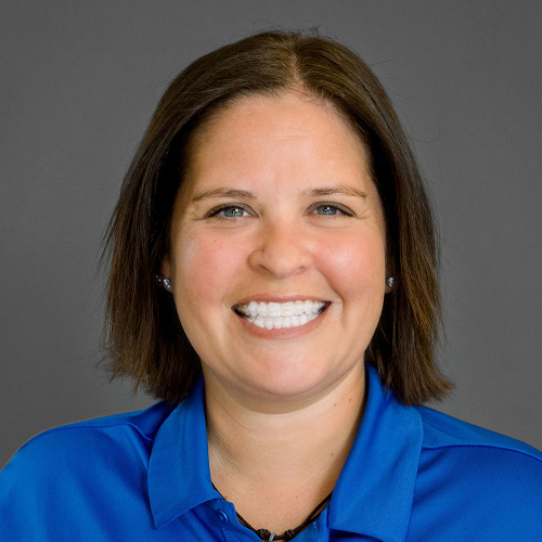 A person with shoulder-length hair wearing a blue collared shirt, with their face blurred out, set against a plain background.