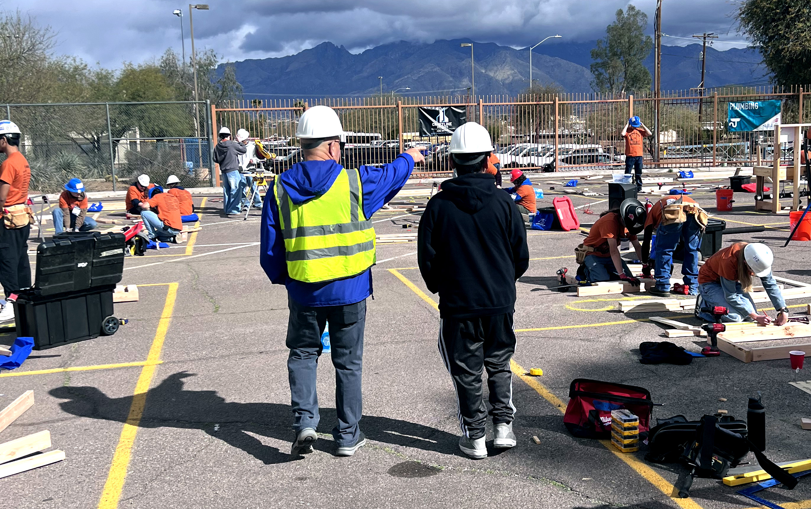 A group of people wearing hard hats work on construction projects in an outdoor area, with two individuals in the foreground observing and pointing.