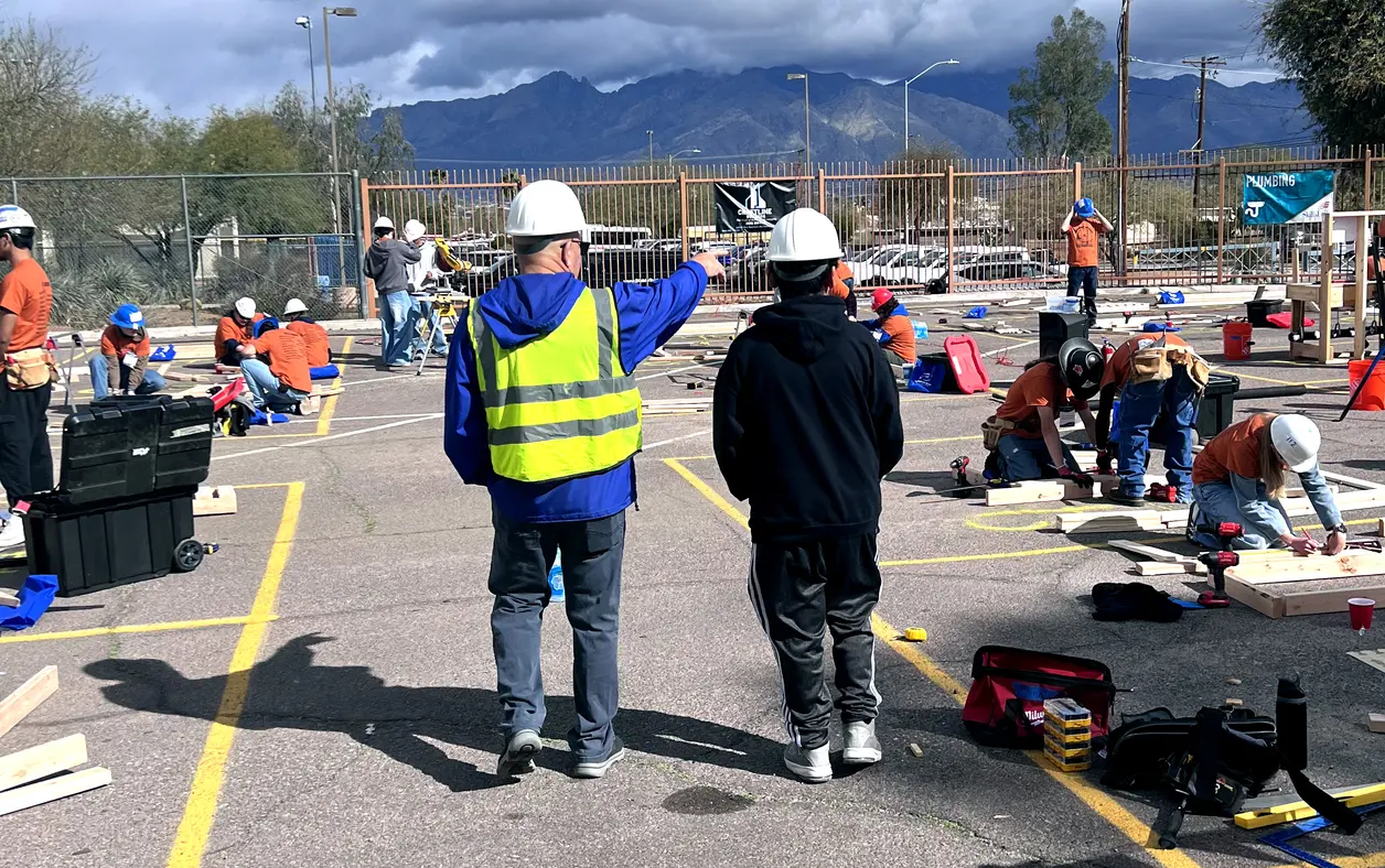 A group of people wearing hard hats work on construction projects in an outdoor area, with two individuals in the foreground observing and pointing. A group of people wearing hard hats work on construction projects in an outdoor area, with two individuals in the foreground observing and pointing.