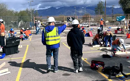A group of people wearing hard hats work on construction projects in an outdoor area, with two individuals in the foreground observing and pointing. A group of people wearing hard hats work on construction projects in an outdoor area, with two individuals in the foreground observing and pointing.