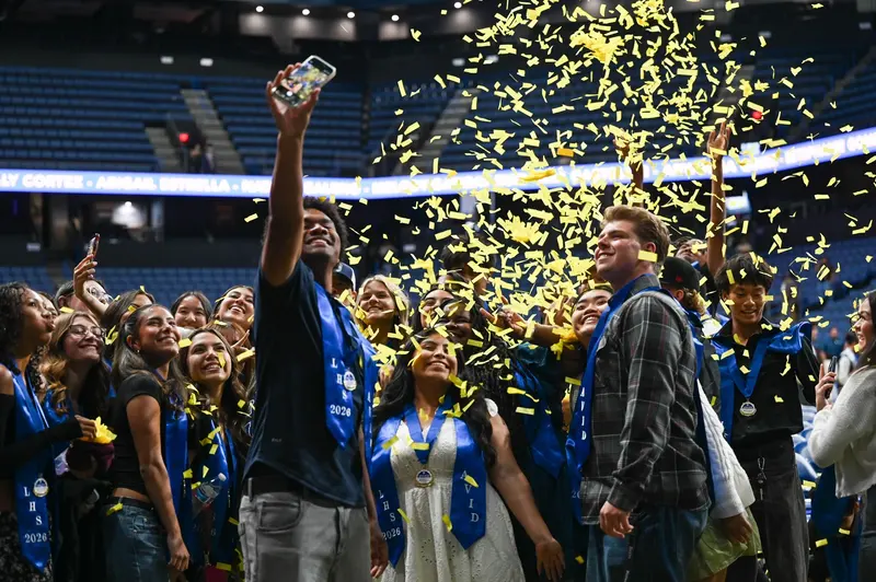 Group of students in graduation attire taking a selfie under falling yellow confetti in an indoor arena. Group of students in graduation attire taking a selfie under falling yellow confetti in an indoor arena.