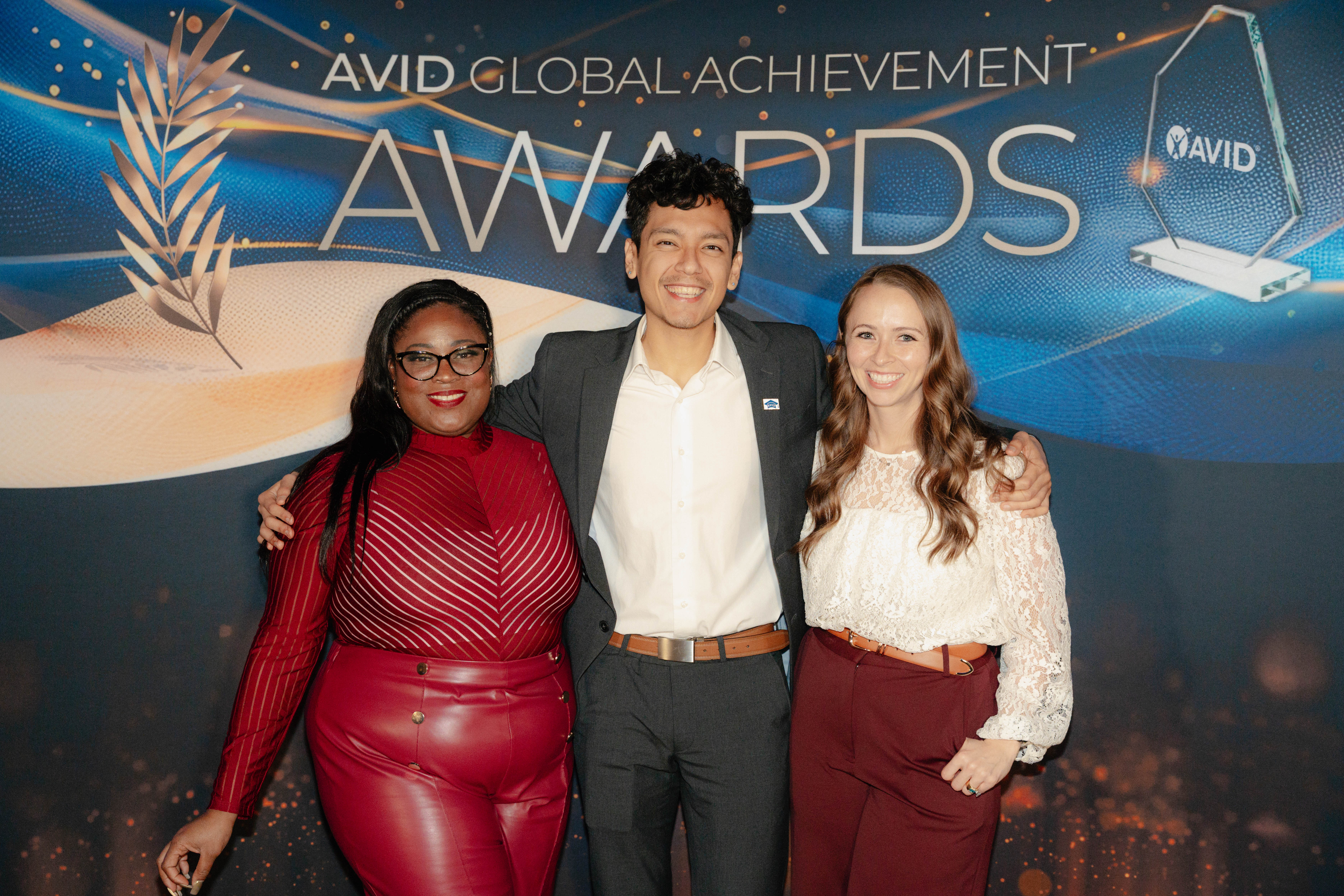 Three people posing together in front of a banner that reads 'AVID Global Achievement Awards'. The banner features decorative elements and an AVID logo on a trophy.