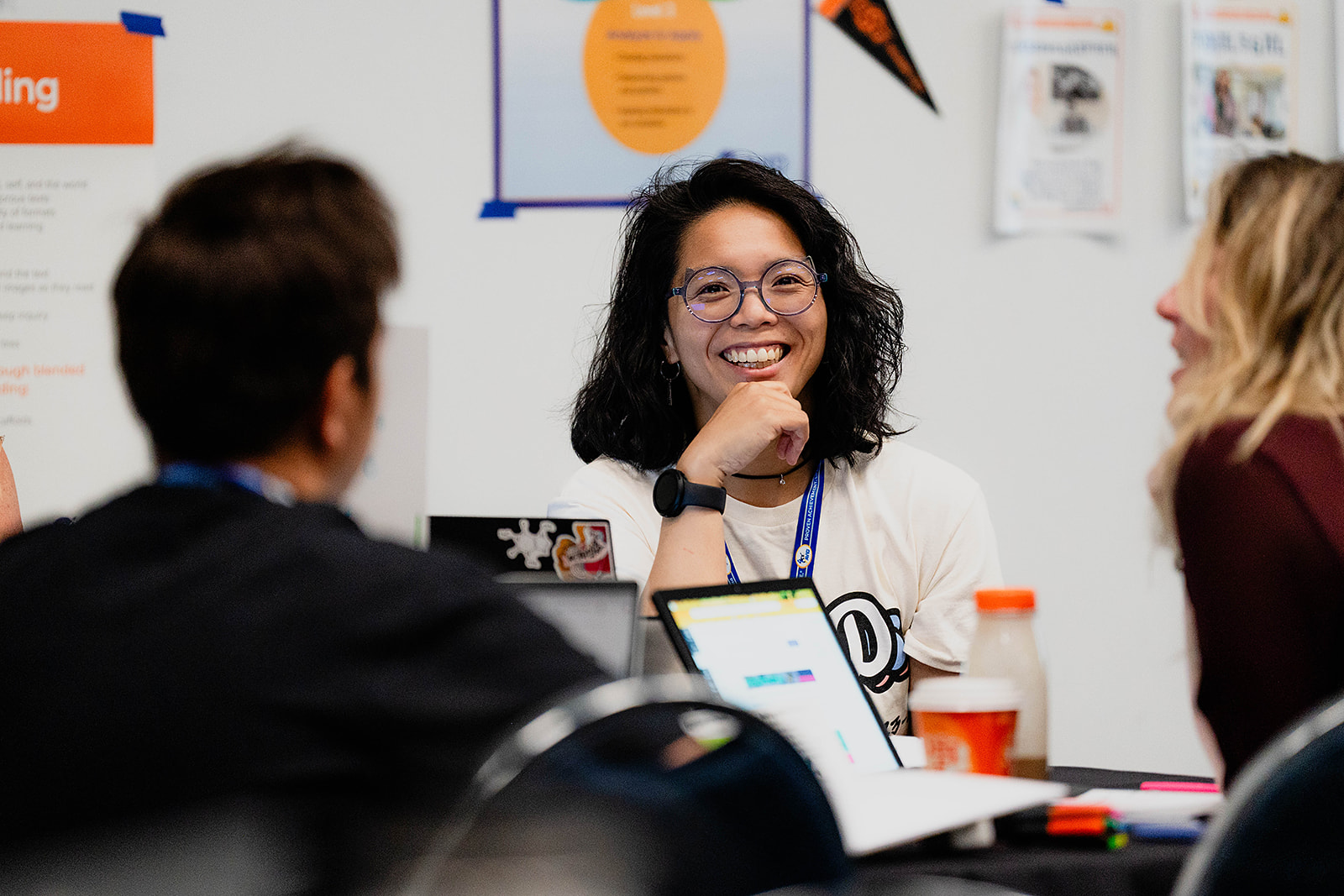 Three individuals sitting at a table with laptops and notebooks, in a classroom or work setting.
