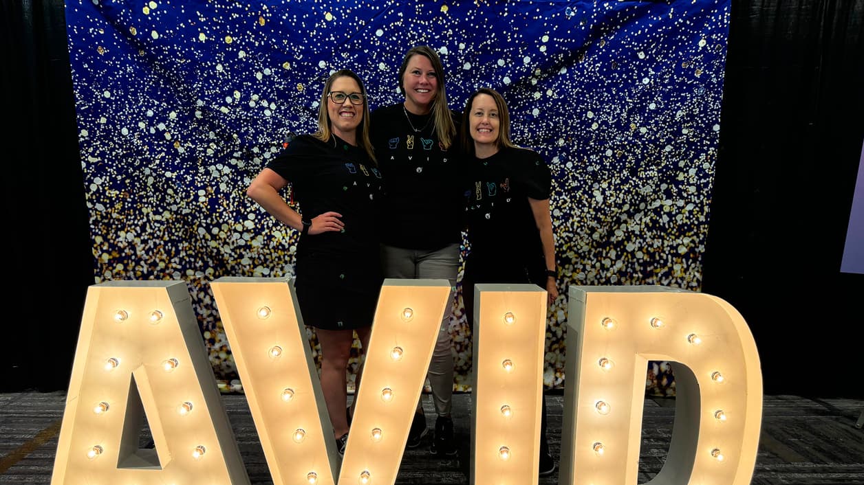 Three people standing in front of a festive, sparkly backdrop and large illuminated letters spelling 'AVID'. Three people standing in front of a festive, sparkly backdrop and large illuminated letters spelling 'AVID'.
