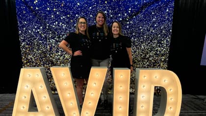 Three people standing in front of a festive, sparkly backdrop and large illuminated letters spelling 'AVID'. Three people standing in front of a festive, sparkly backdrop and large illuminated letters spelling 'AVID'.