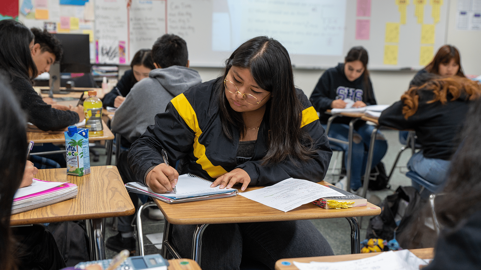 Students in a classroom working on assignments, one student is prominently focused on writing in a notebook at a desk.