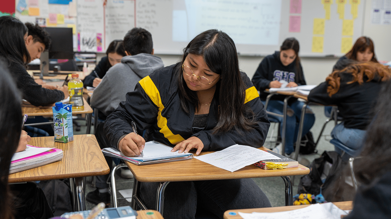Students in a classroom working on assignments, one student is prominently focused on writing in a notebook at a desk. Students in a classroom working on assignments, one student is prominently focused on writing in a notebook at a desk.