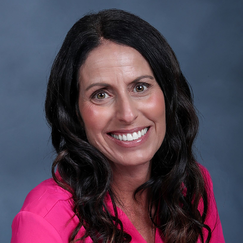 Individual with long dark hair wearing a bright pink shirt against a dark blue background