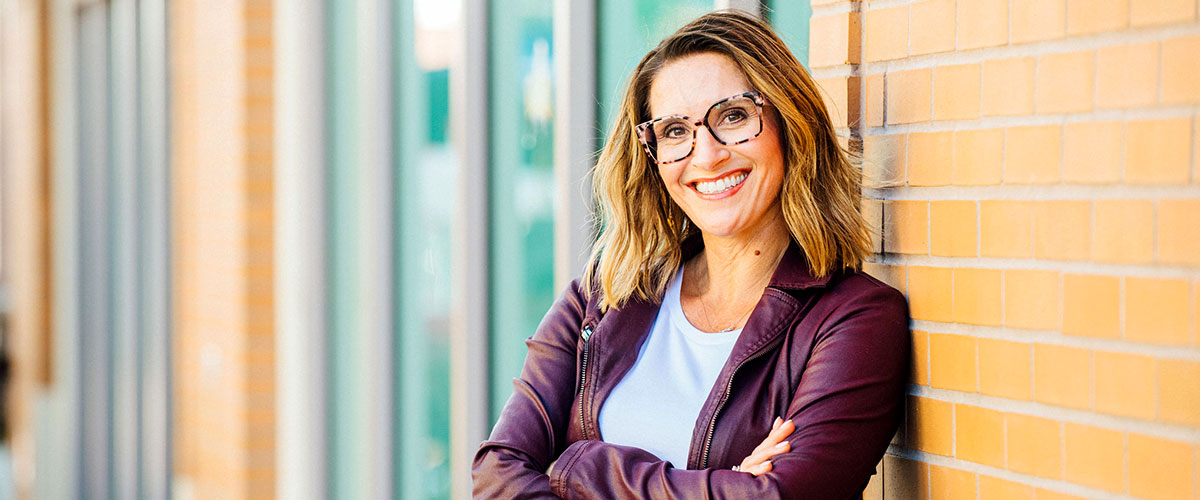 Person leaning against a brick wall with crossed arms wearing a purple jacket and white shirt.