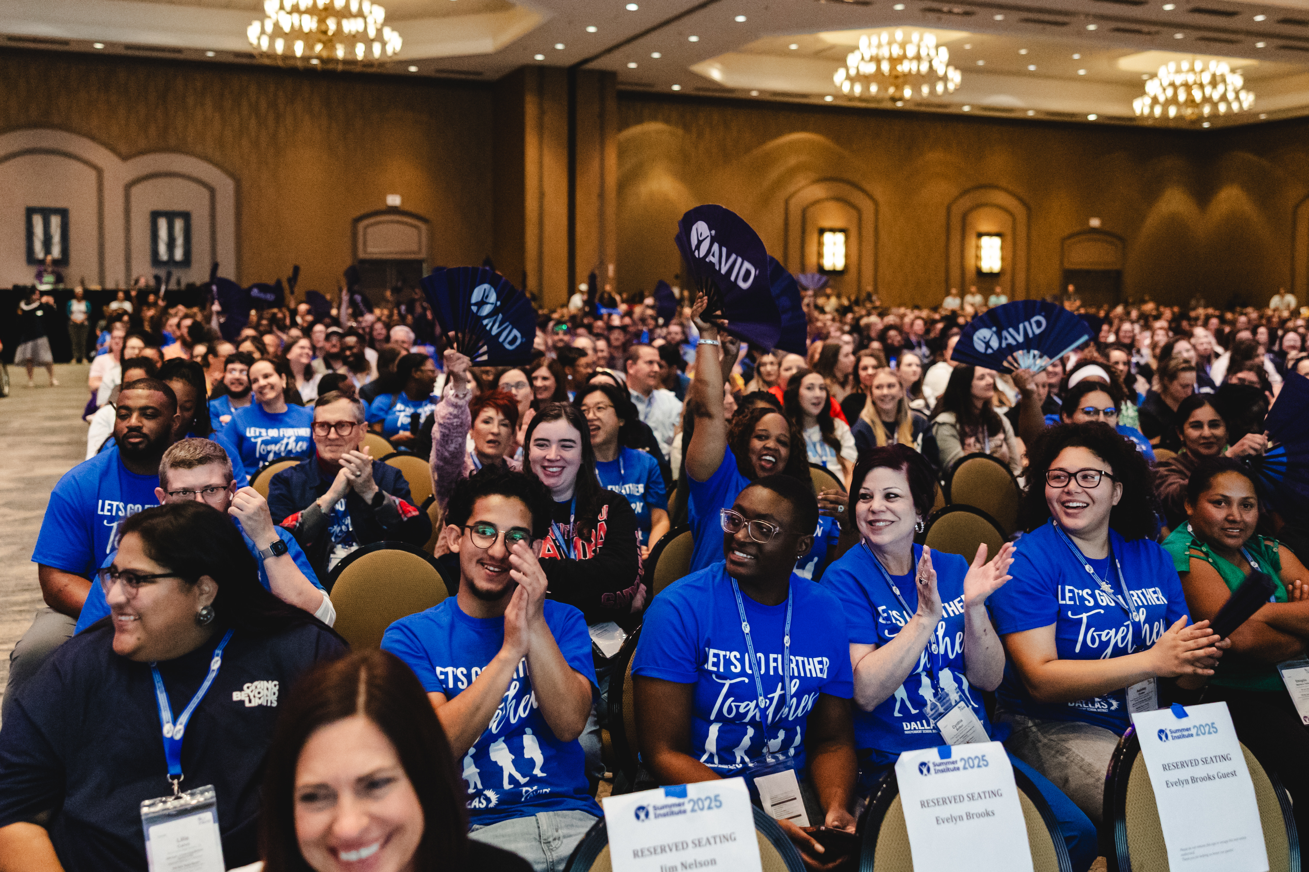 A large crowd of people seated in a conference room, wearing blue shirts and holding AVID signs.