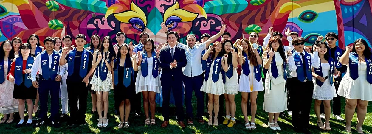 A large group of people wearing graduation stoles, posing in front of a colorful mural. A large group of people wearing graduation stoles, posing in front of a colorful mural.