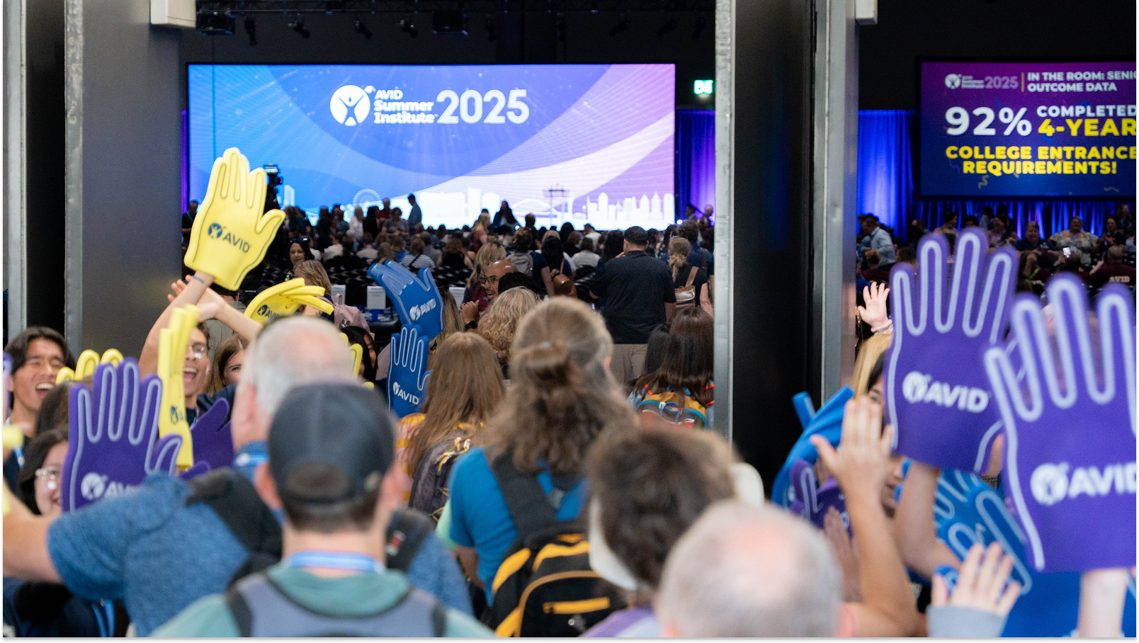 Attendees at a conference holding AVID foam hands in a crowded event hall with a large screen displaying 'AVID Summer Institute 2025' in the background.