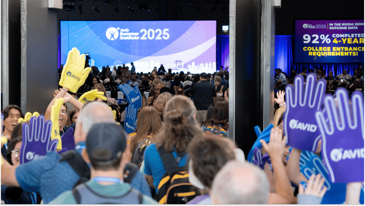 Attendees at a conference holding AVID foam hands in a crowded event hall with a large screen displaying 'AVID Summer Institute 2025' in the background. Attendees at a conference holding AVID foam hands in a crowded event hall with a large screen displaying 'AVID Summer Institute 2025' in the background.