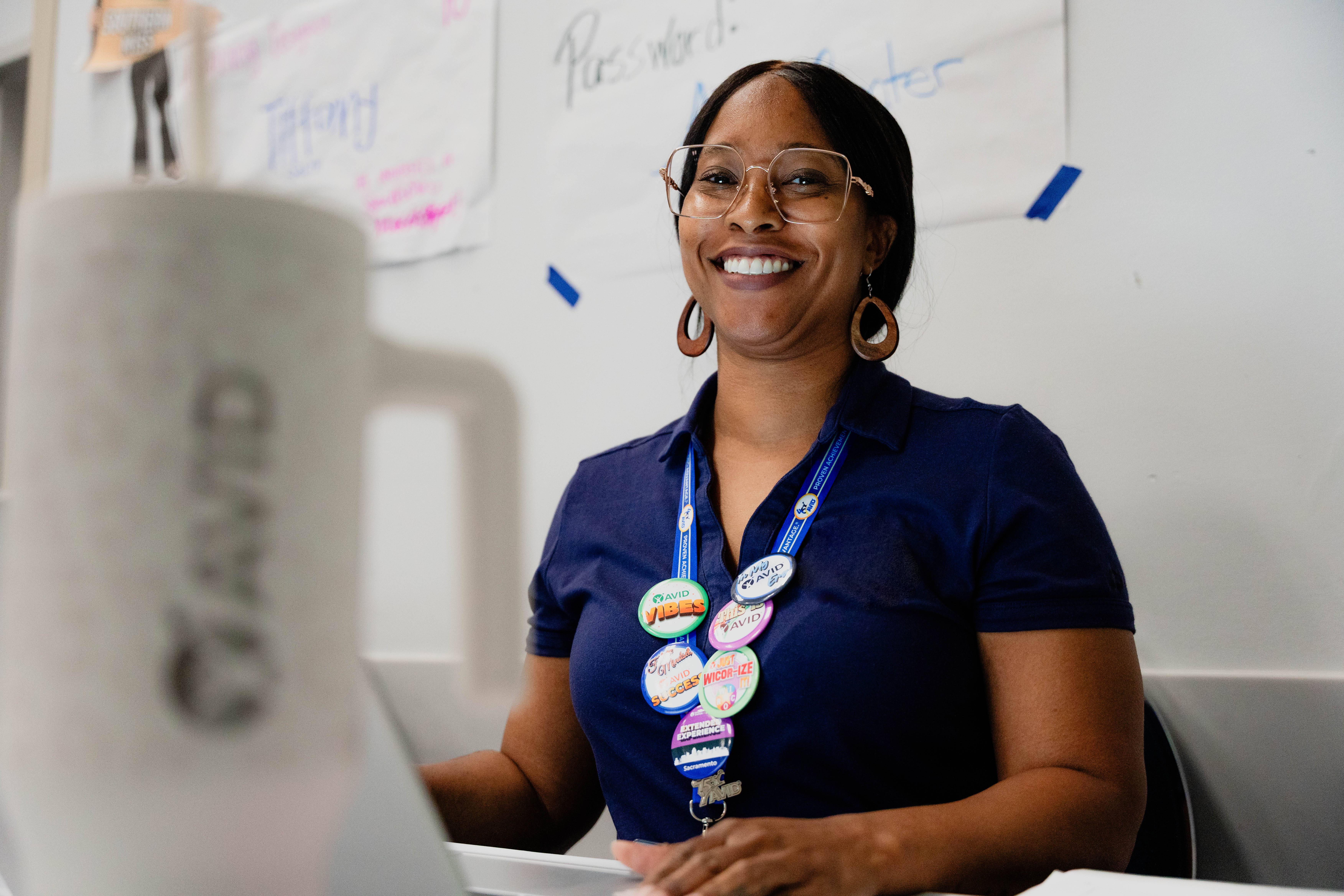 Person wearing a dark blue shirt and a lanyard with multiple colorful buttons sits at a desk with posters in the background.