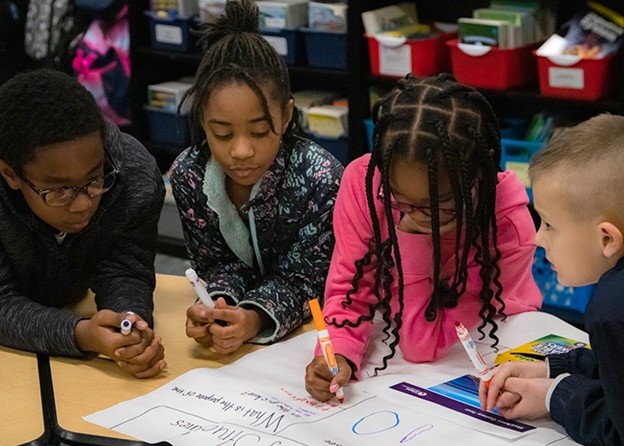 Children collaborating and drawing together at a table in a colorful classroom setting.