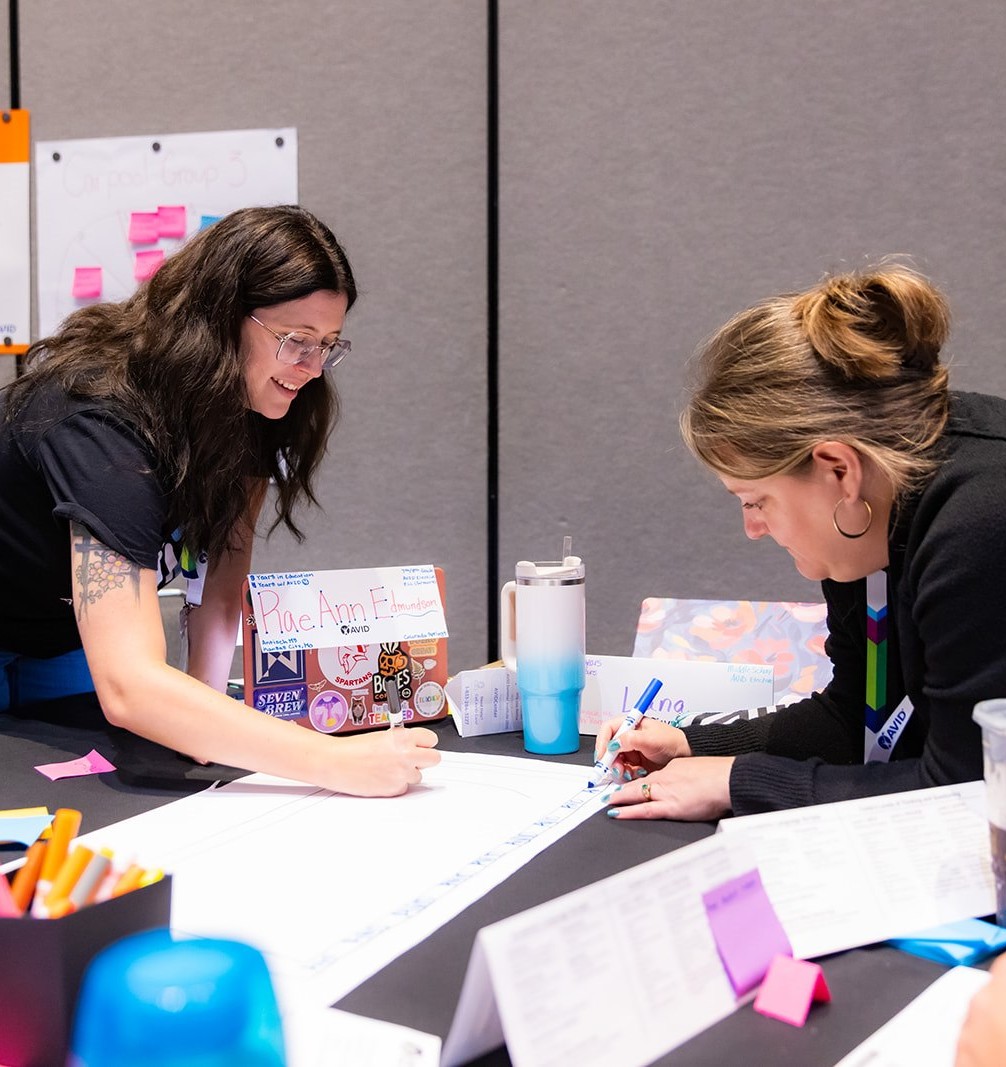 Two individuals working on a project at a table, writing on a large paper with various supplies around them.