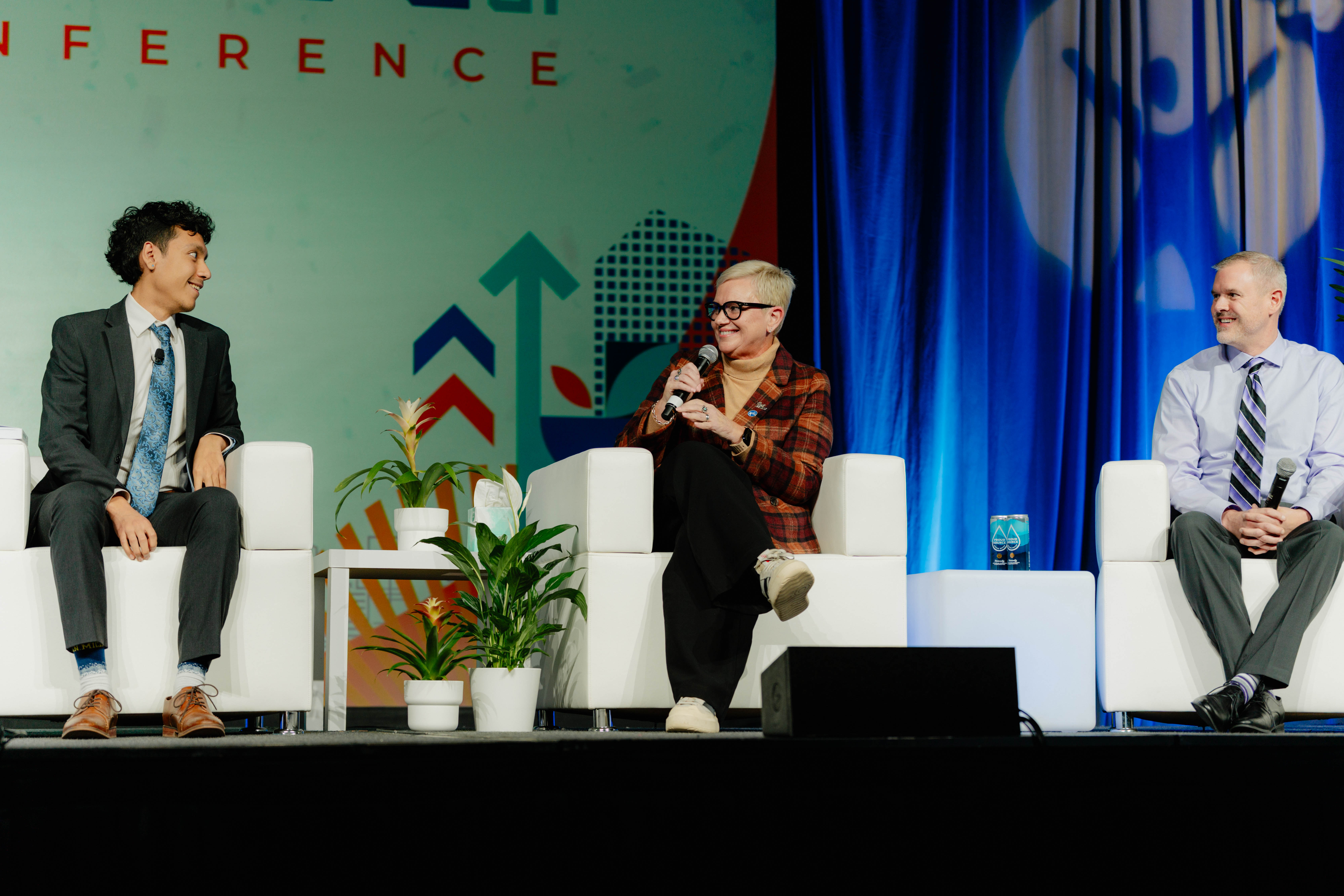 Three individuals seated on stage at a conference, engaged in a conversation with microphones. There are plants on the table between them, and a colorful background with arrows and patterns.