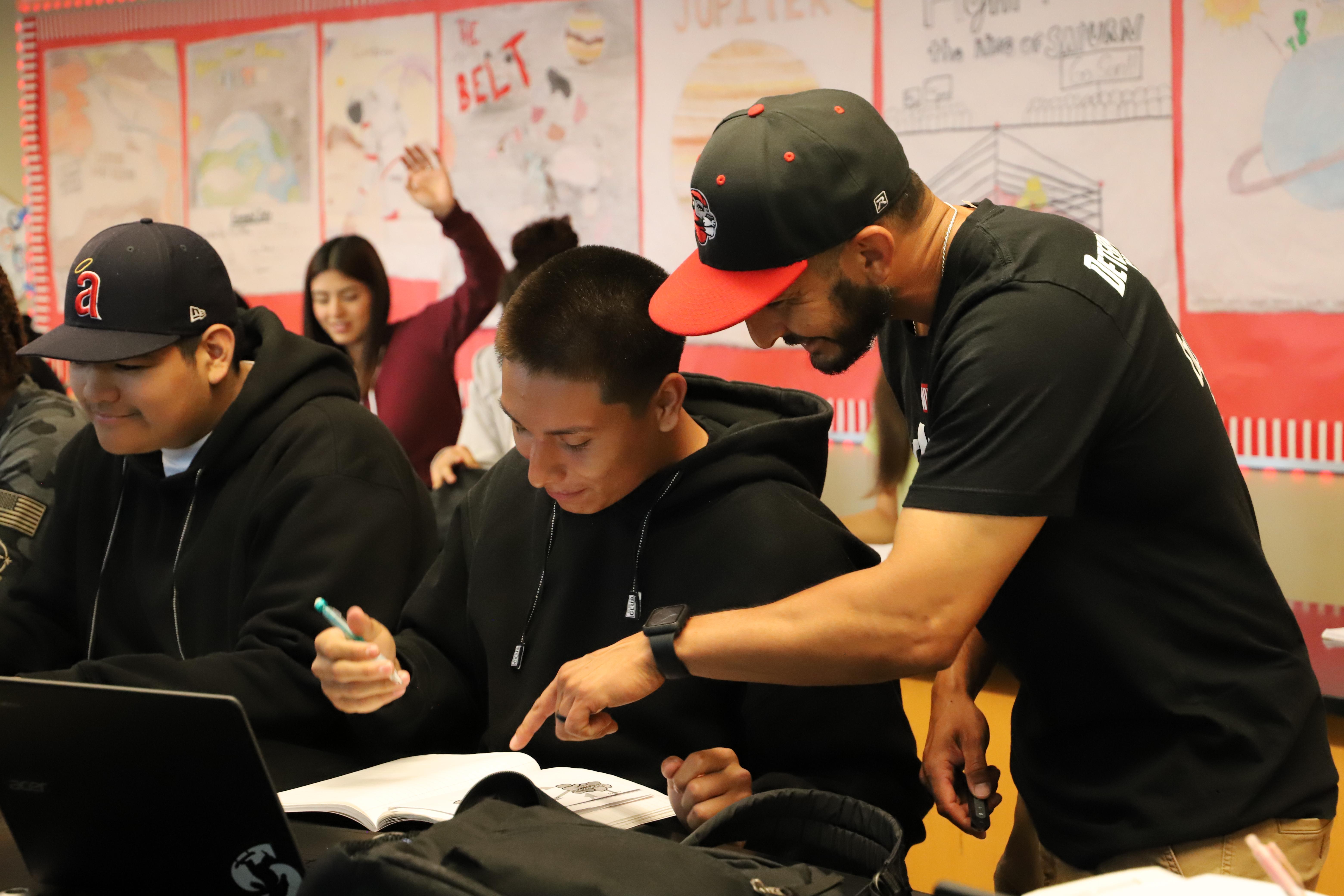 A classroom scene with a teacher assisting a student, pointing at a textbook on a desk. Other students are seen in the background with educational posters on the wall.