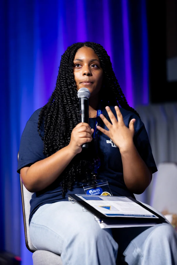 Person seated on a chair holding a microphone with a lanyard and a clipboard on their lap, in front of a blue and purple backdrop.