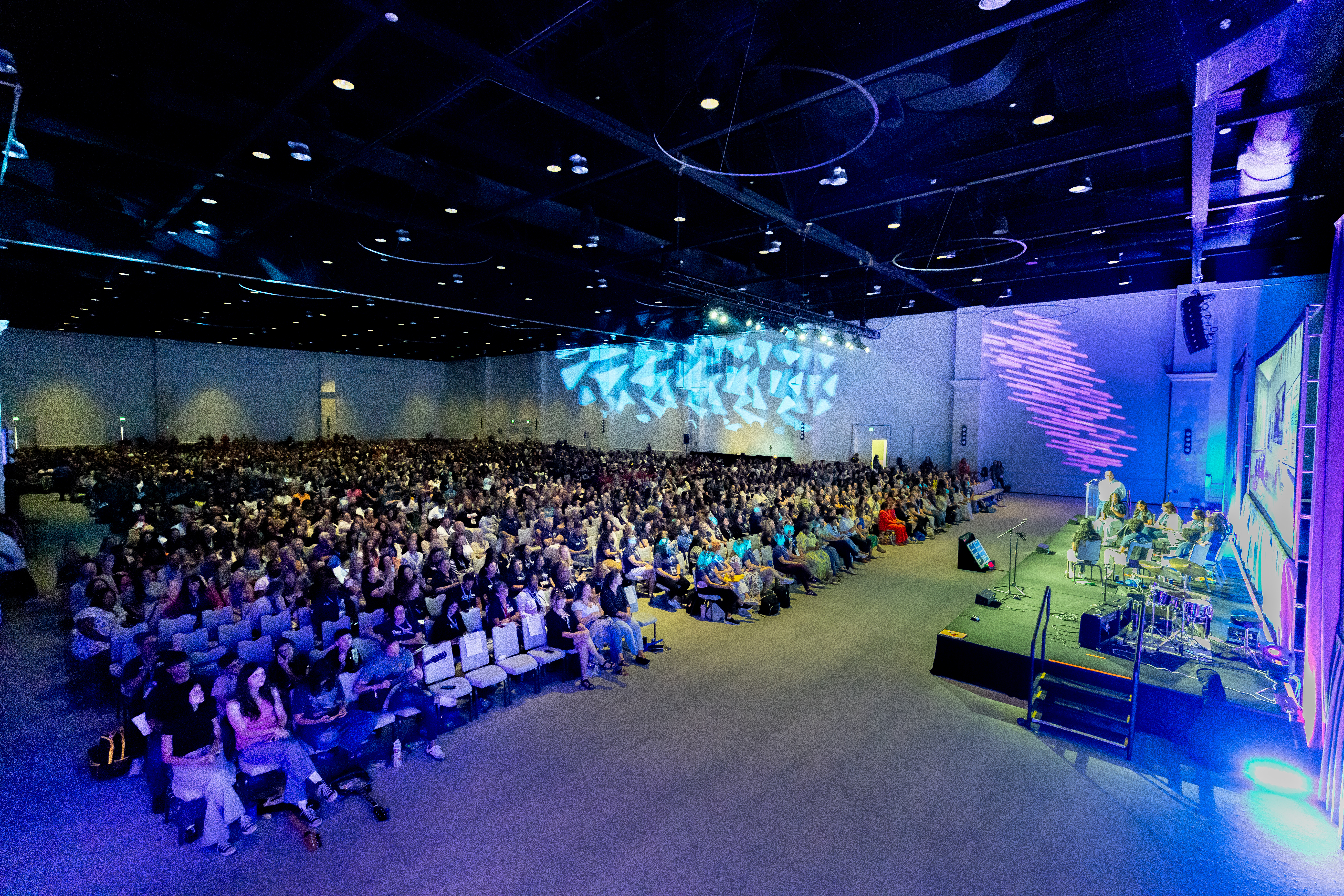A wide-angle view of a large indoor event with a stage and an audience seated in rows, under dim lighting with blue and purple hues.