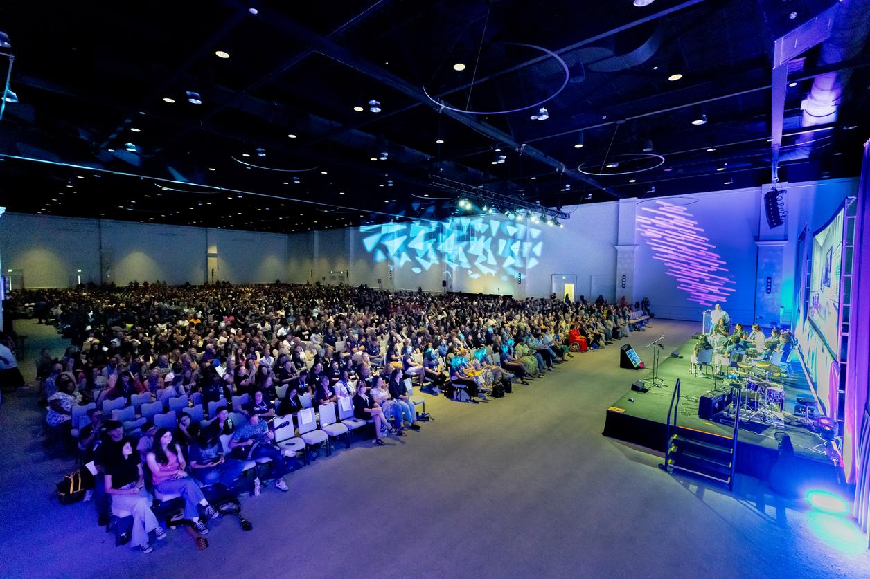 A wide-angle view of a large indoor event with a stage and an audience seated in rows, under dim lighting with blue and purple hues. A wide-angle view of a large indoor event with a stage and an audience seated in rows, under dim lighting with blue and purple hues.
