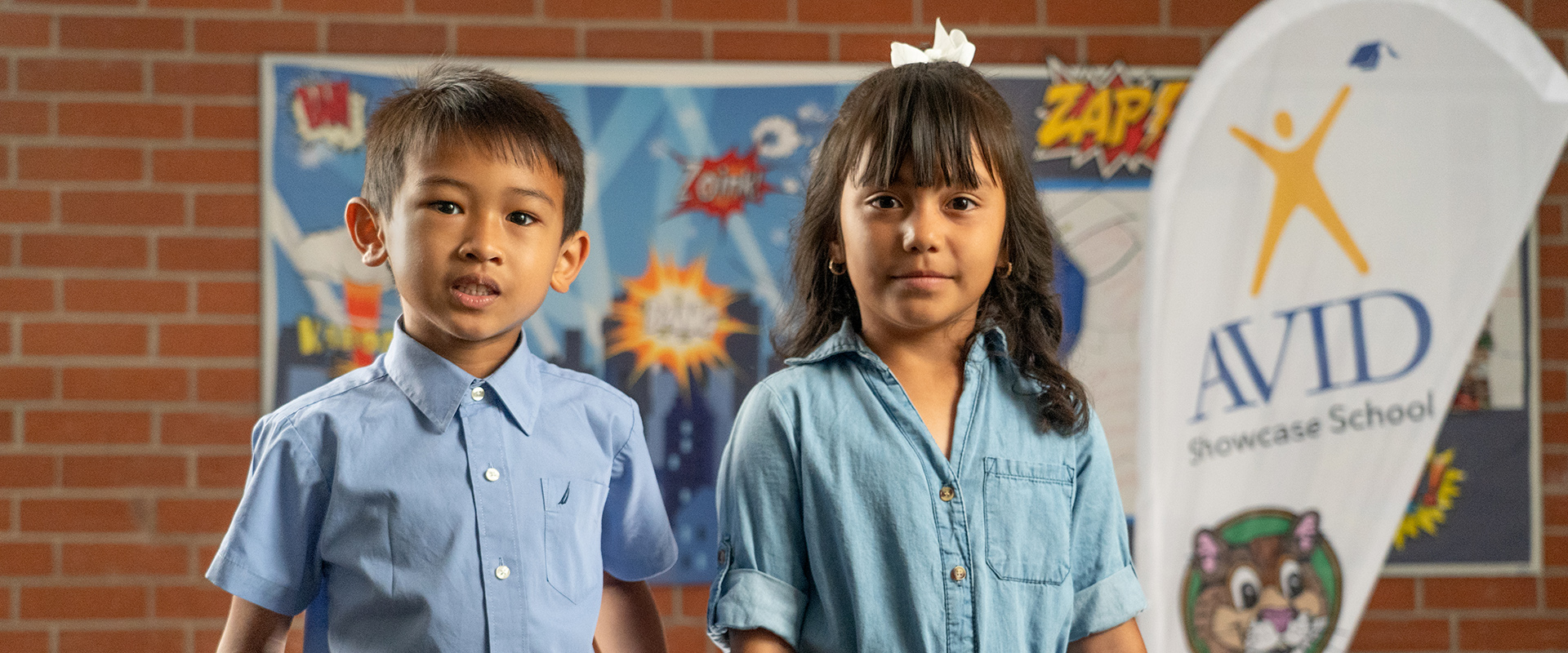 Two children in blue shirts standing in front of a colorful background featuring comic-style art and an AVID Showcase School banner.