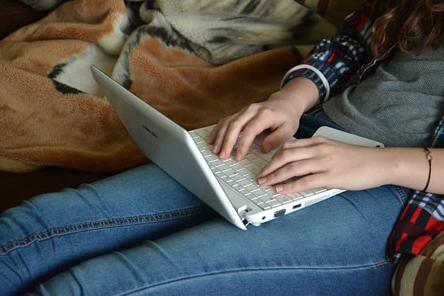 A person sitting on a couch, typing on a white laptop placed on their lap. The person's hands are visible on the keyboard, with a blanket in the background. A person sitting on a couch, typing on a white laptop placed on their lap. The person's hands are visible on the keyboard, with a blanket in the background.