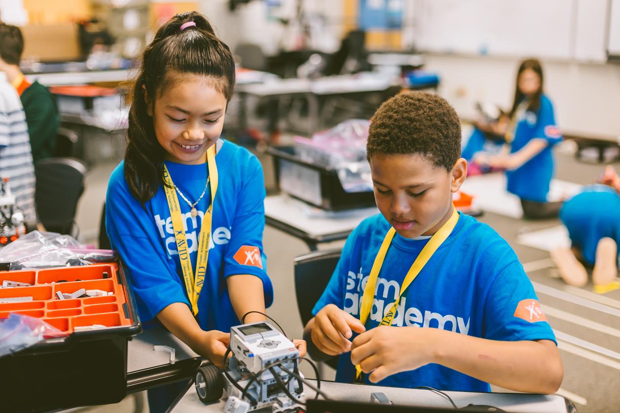 Two children in blue shirts working on a robotics project in a classroom setting with other children in the background. Two children in blue shirts working on a robotics project in a classroom setting with other children in the background.