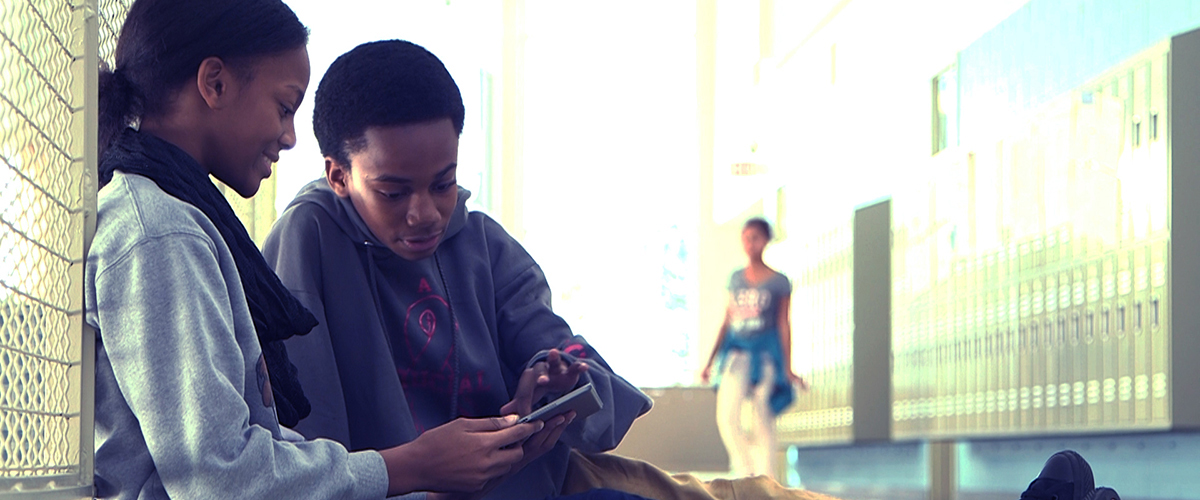 Two individuals sitting next to a chain-link fence, one using a tablet, with lockers and a passerby in the background.