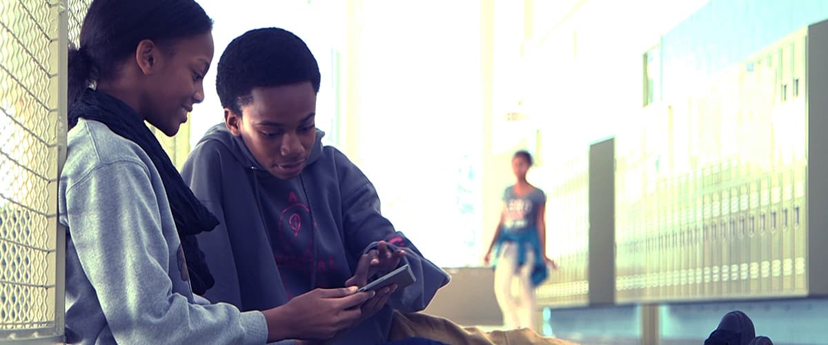 Two individuals sitting next to a chain-link fence, one using a tablet, with lockers and a passerby in the background. Two individuals sitting next to a chain-link fence, one using a tablet, with lockers and a passerby in the background.