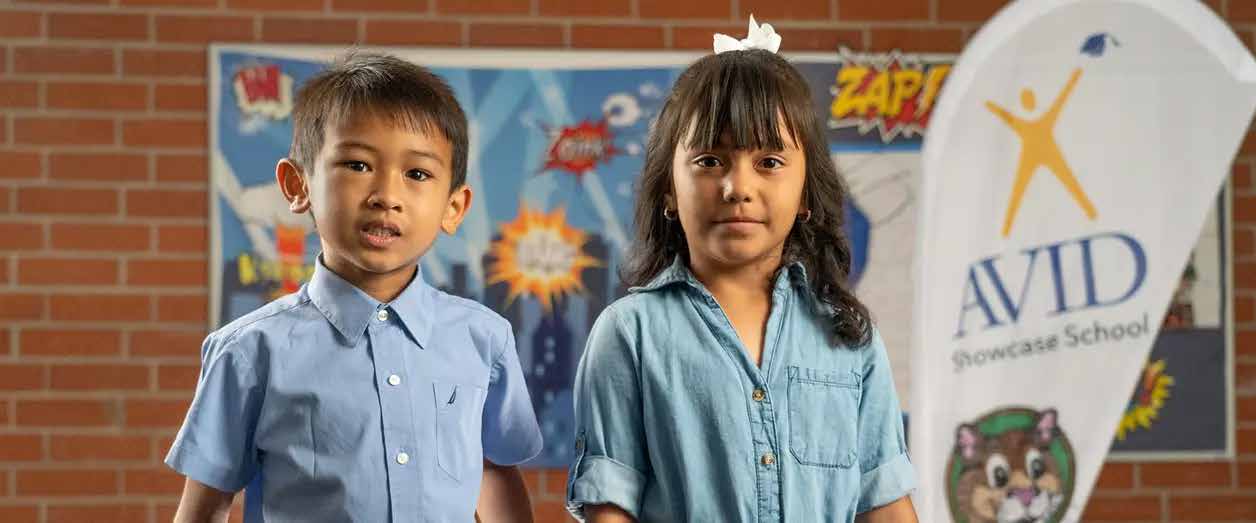 Two children standing side by side in front of a brick wall with a school banner and comic-style artwork in the background.