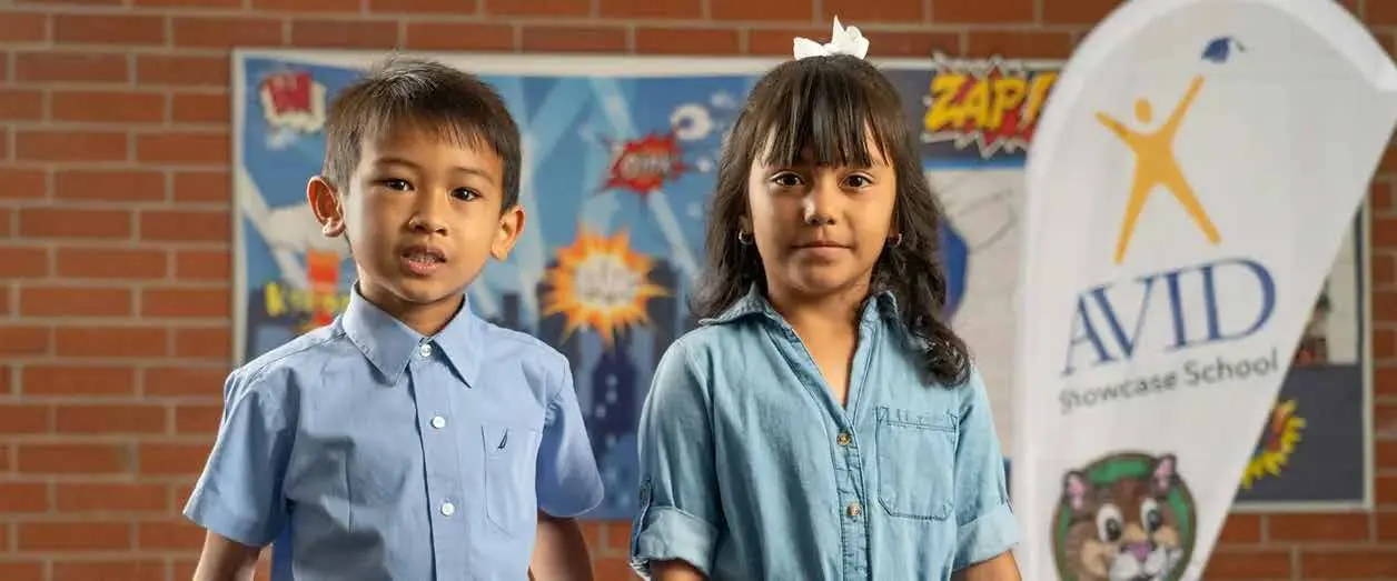 Two children standing side by side in front of a brick wall with a school banner and comic-style artwork in the background. Two children standing side by side in front of a brick wall with a school banner and comic-style artwork in the background.